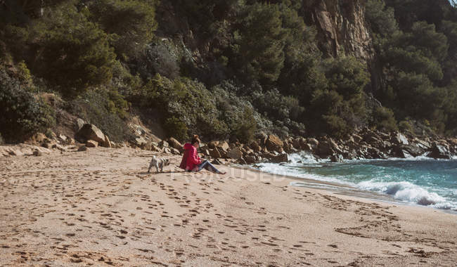 Зріла жінка в супроводі мопса собаки читає книгу на березі моря в сонячний день — стокове фото
