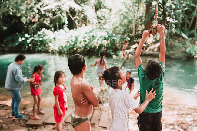 LAOS, LUANG PRABANG: Crianças brincando com baloiço de corda em pequena lagoa na floresta ensolarada . — Fotografia de Stock