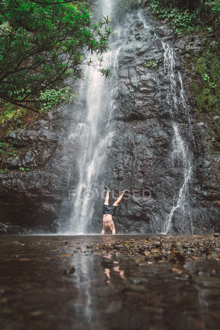 Akrobat-Mann steht auf Händen neben Wasserfall. — Stockfoto