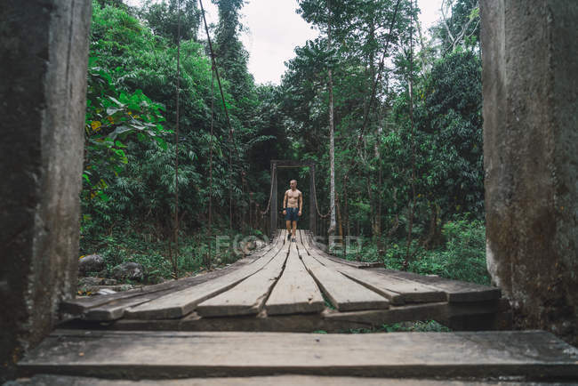 Bello uomo in forma senza maglietta che cammina su un ponte di legno nella foresta . — Foto stock