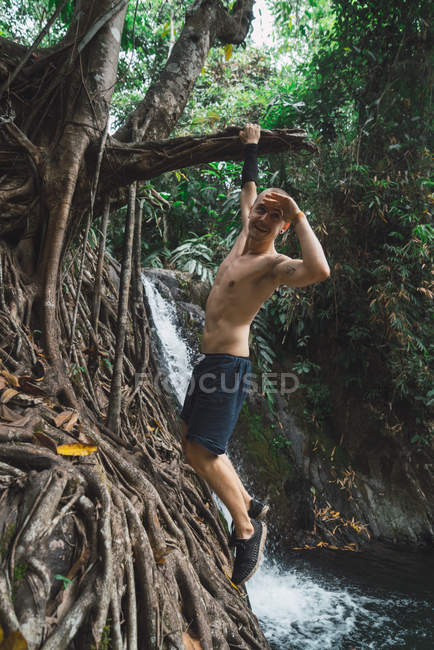 Cheerful acrobatic man hanging on tree and looking at camera with hand up to head. — Stock Photo