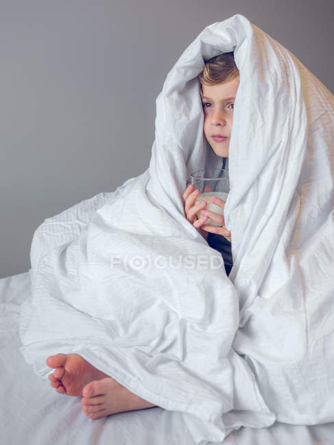 Netter kleiner Junge in Bettdecke gewickelt sitzt und trinkt ein Glas Milch. — Stockfoto