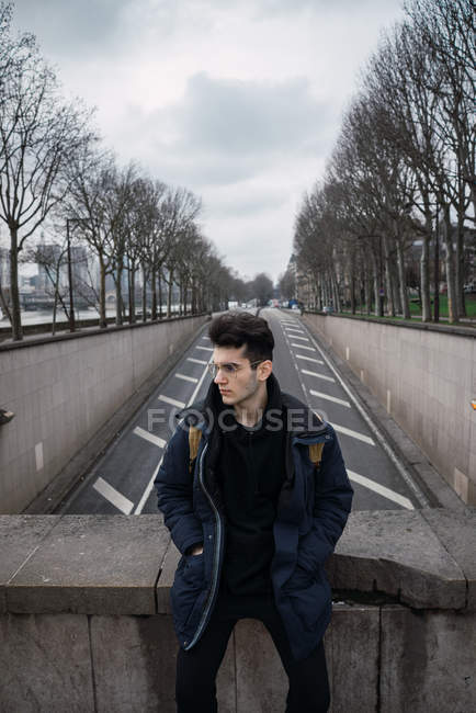Продуманий молодий чоловік в окулярах сидить на фоні асфальтової дороги в місті . — стокове фото