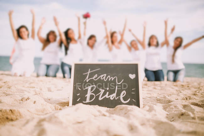 Pizarra con letras de novia de equipo sobre mujeres irreconocibles posando con las manos en la playa . - foto de stock