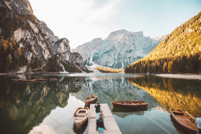Personnes méconnaissables marchant sur une jetée en bois avec des bateaux au lac dans les montagnes . — Photo de stock