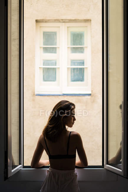Visão traseira da menina posando pela janela em casa e olhando sobre o ombro na câmera — Fotografia de Stock