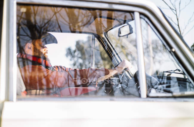 Vista lateral do homem barbudo em chapéu dirigindo carro branco na natureza . — Fotografia de Stock