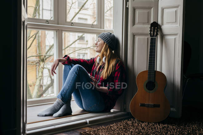 Улыбающаяся молодая женщина, сидящая у окна на гитаре — стоковое фото