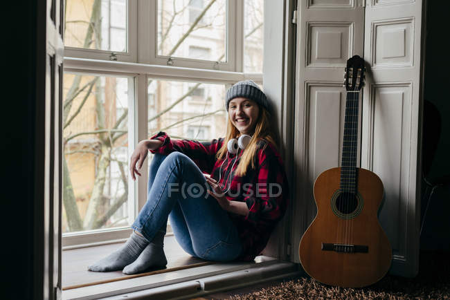 Smiling young blonde  sitting on window sill by guitar and looking at camera. — Stock Photo