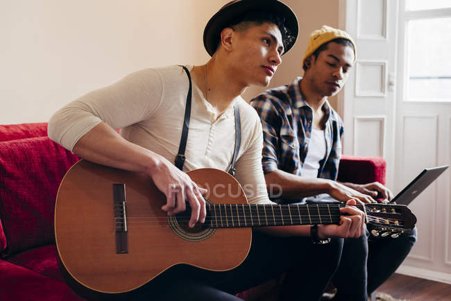 Молодые стильные мужчины отдыхают на диване с ноутбуком и гитарой — стоковое фото