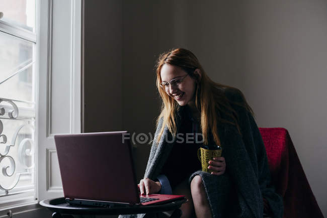 Frau im Sessel blättert im Laptop und trinkt Kaffee — Stockfoto