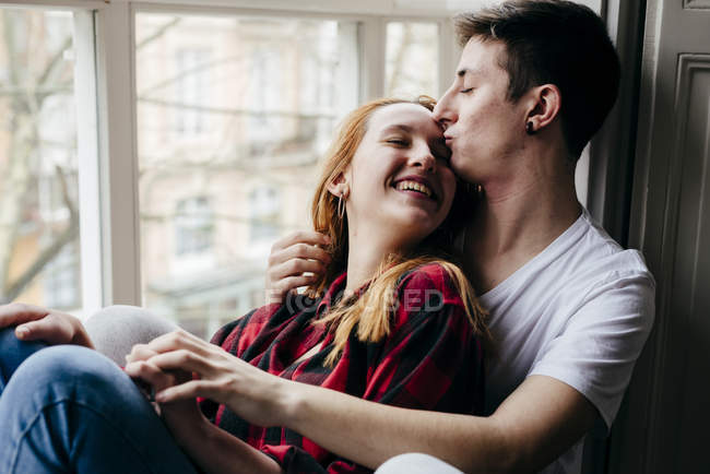 Young man kissing blonde girlfriend in foreground at window — Stock Photo