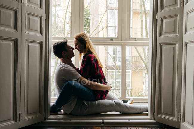 Alegre pareja sentado en ventana alféizar y abrazar - foto de stock