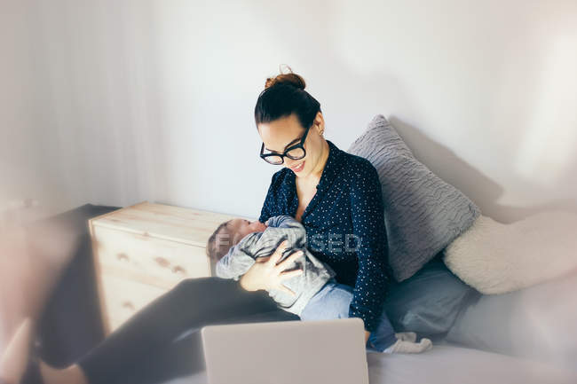 Улыбающаяся женщина держит спящего ребенка на руках и просматривает ноутбук — стоковое фото