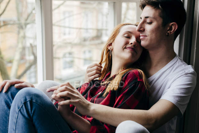 Sensual joven pareja abrazándose en ventana alféizar con los ojos cerrados - foto de stock