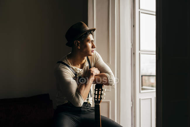 Homme réfléchi en chapeau appuyé sur la guitare et regardant la fenêtre — Photo de stock