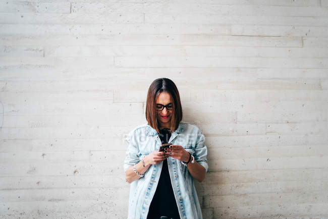 Young woman standing at concrete wall and browsing smartphone. — Stock Photo