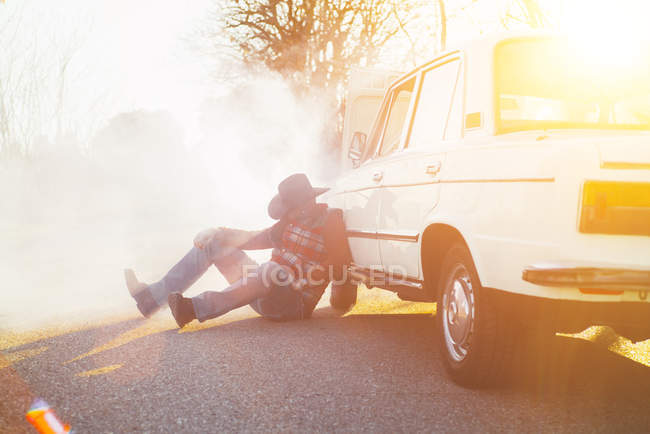 Hombre con sombrero apoyado en coche roto emitiendo humo en la carretera . - foto de stock