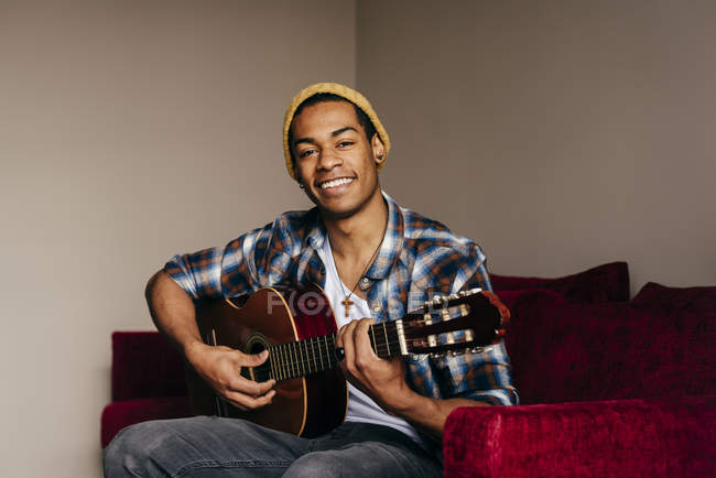 Jovem alegre sentado no sofá e tocando guitarra acústica em casa . — Fotografia de Stock