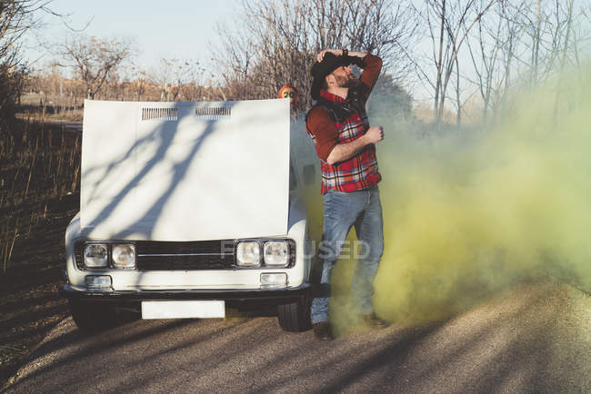 Man in hat by broken vintage car emitting smoke in nature. — Stock Photo