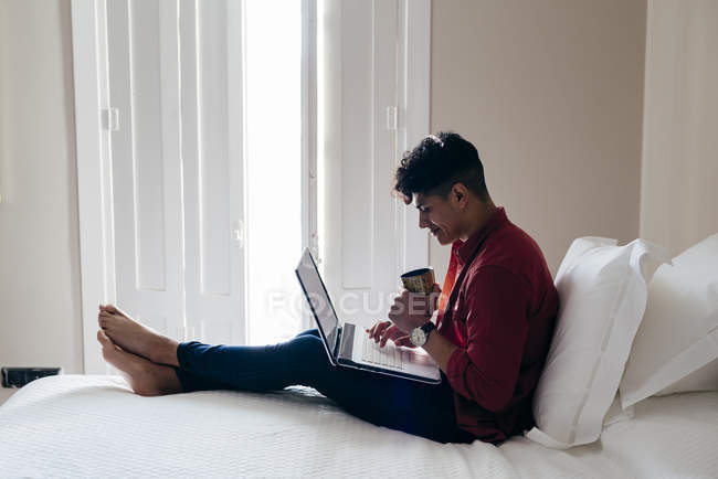 Homem com copo sentado na cama e laptop de navegação — Fotografia de Stock