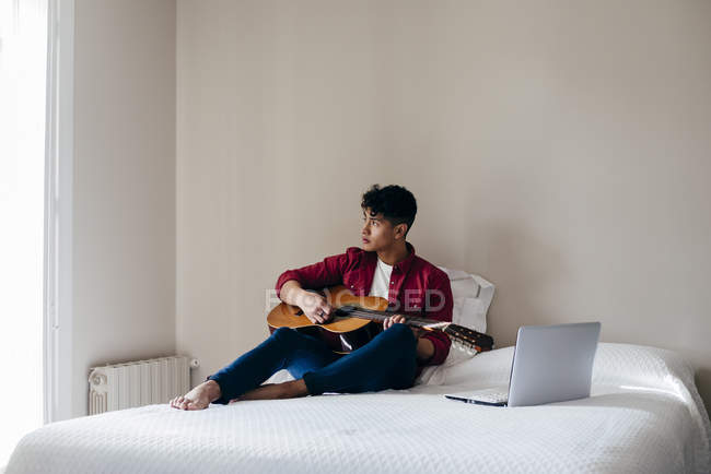 Homme avec guitare assis sur le lit et regardant ailleurs — Photo de stock