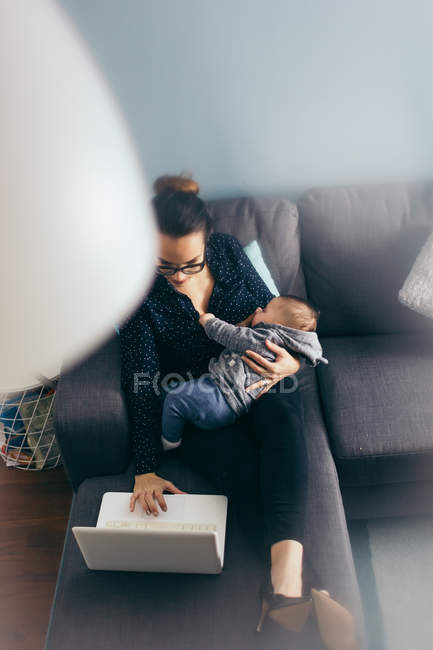 Smiling woman sitting on coach with sleeping child and browsing laptop — Stock Photo