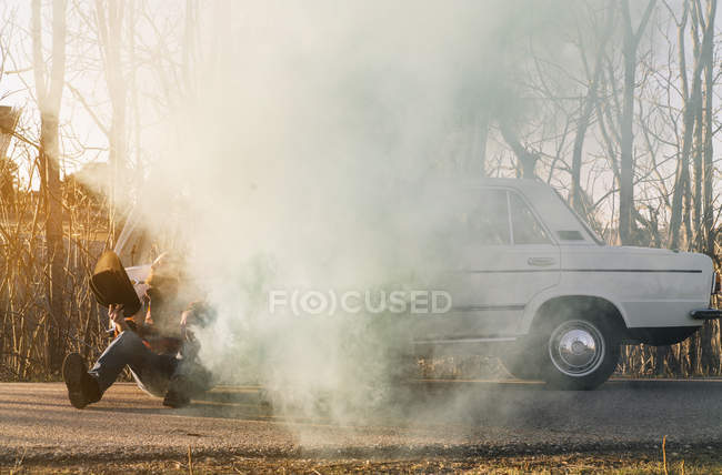 Человек в шляпе опирается на сломанную винтажную машину, излучающую дым, припаркованный на обочине дороги. . — стоковое фото