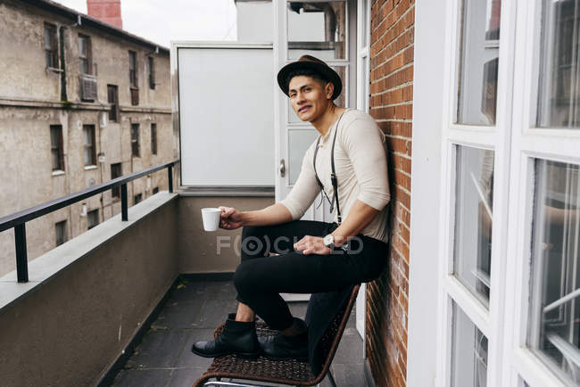 Man in hat drinking coffee on balcony and looking at camera — Stock Photo