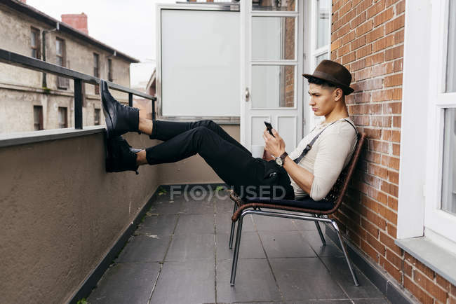 Homem de roupas vintage sentado na varanda e smartphone de navegação — Fotografia de Stock