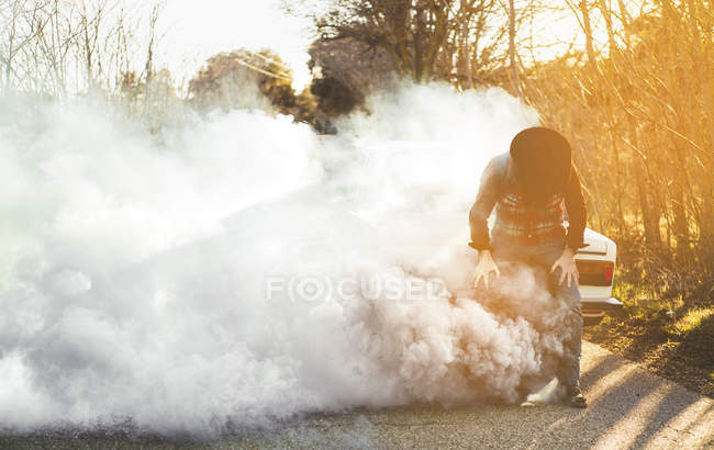 Unrecognizable tired man in hat leaning on vintage car emitting smoke in countryside. — Stock Photo