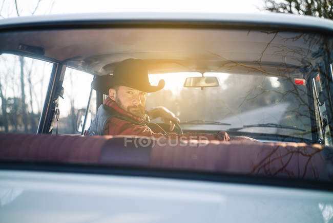 Hombre en sombrero de conducción vintage coche blanco en la naturaleza y mirando hacia atrás por encima del hombro - foto de stock