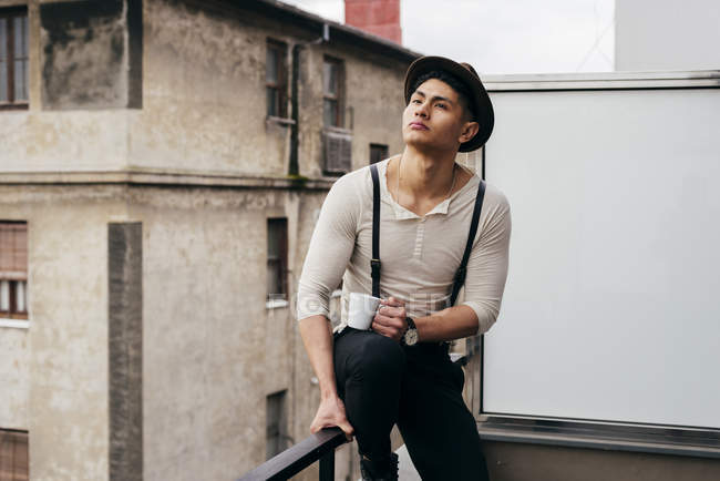 Stilvoller Mann mit Tasse sitzt auf Balkongeländer und schaut weg — Stockfoto