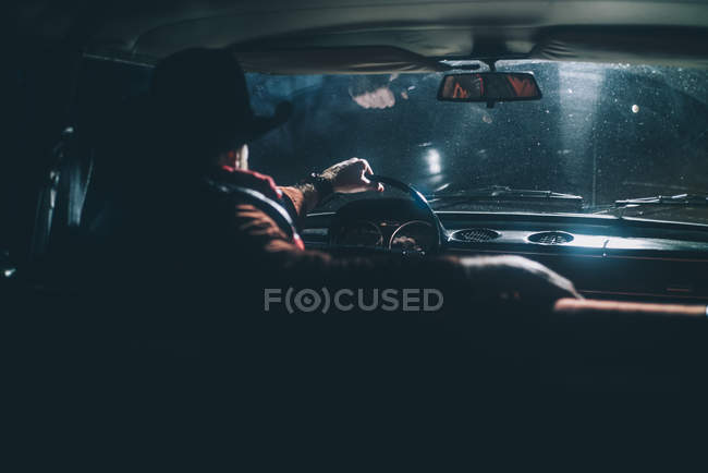 Rear view of man in hat driving car with headlights on at night. — Stock Photo