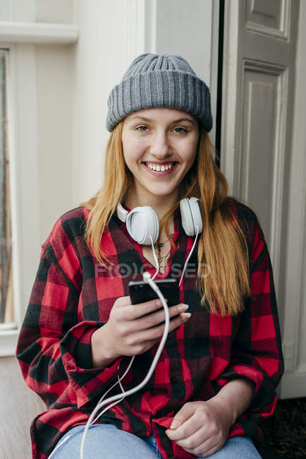 Усміхнена молода блондинка з навушниками і смартфоном і дивиться на камеру . — стокове фото