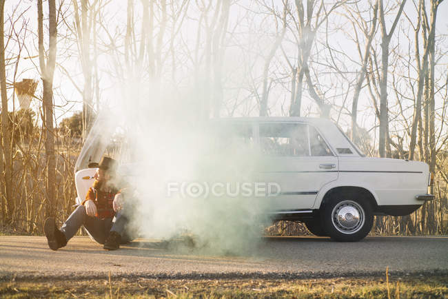 Человек в шляпе опирается на разбитую машину, излучающую дым в сельской местности — стоковое фото