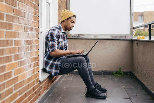 Seitenansicht eines stilvollen Mannes, der auf dem Balkon sitzt und auf dem Laptop tippt. — Stockfoto