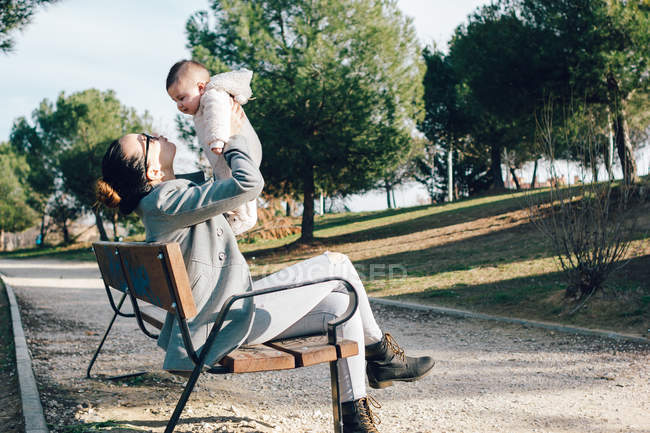 Mujer alegre sentada en el banco del parque y sosteniendo al niño en el aire - foto de stock