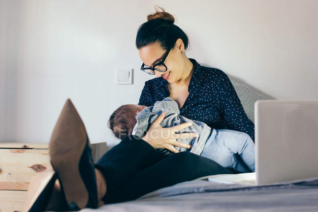 Lächelnde Frau mit Brille sitzt mit Laptop im Bett und hält schlafenden Sohn. — Stockfoto