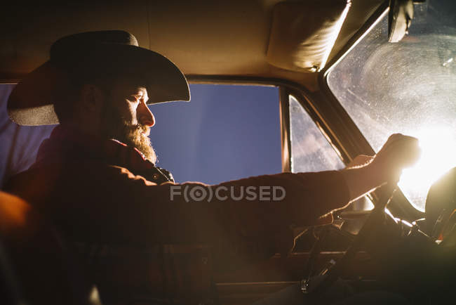 Vista lateral del hombre en el coche de la conducción del sombrero en la luz del atardecer - foto de stock