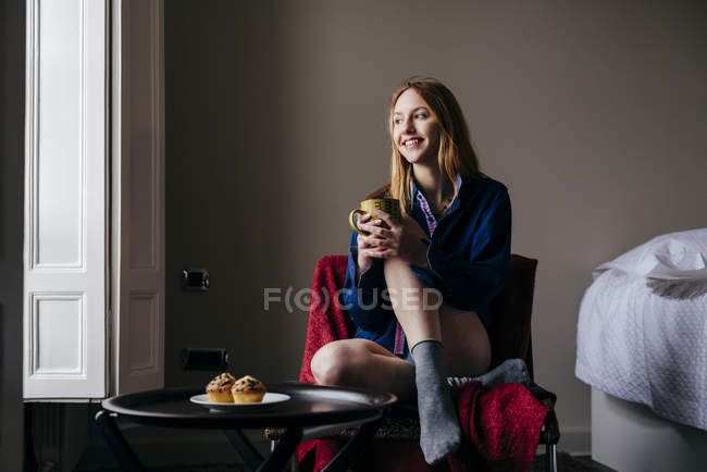 Mujer rubia sonriente sentada en sillón con taza en casa - foto de stock