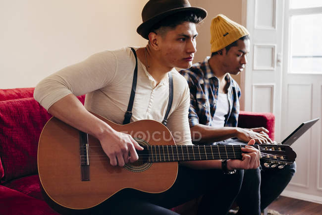 Молодий стильний чоловік грає на гітарі, коли друг використовує ноутбук на дивані вдома . — стокове фото