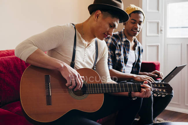 Young man playing guitar while friend using laptop on sofa at home. — Stock Photo