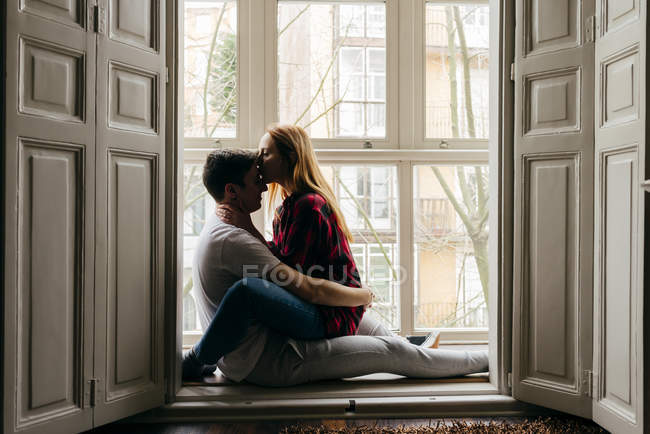 Vista lateral de pareja joven besándose en el alféizar de la ventana en casa - foto de stock