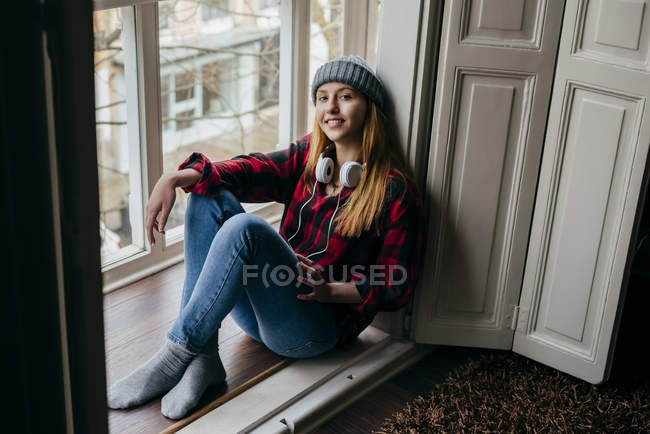 Улыбающаяся молодая женщина в наушниках сидит на подоконнике и смотрит в камеру — стоковое фото