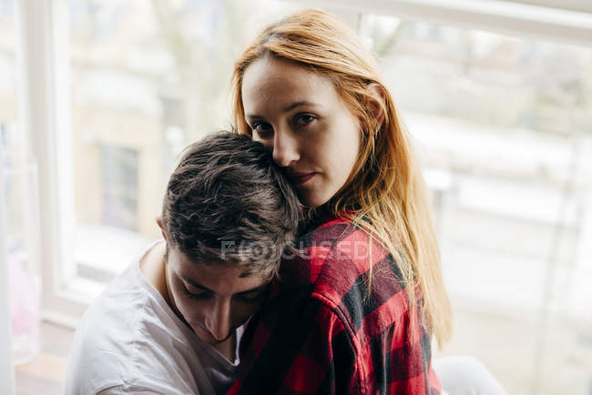 Young blonde girl embracing with boyfriend at window and looking at camera — Stock Photo