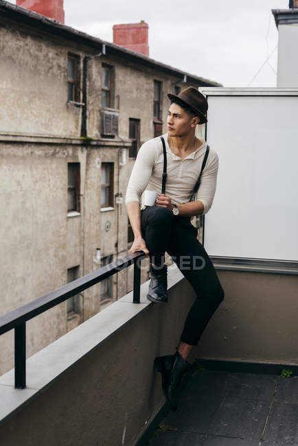 Elegante joven con sombrero sentado con taza en el balcón y mirando hacia otro lado - foto de stock