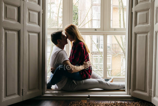 Vista lateral do jovem e da mulher sentados juntos e beijando no peitoril da janela em casa . — Fotografia de Stock