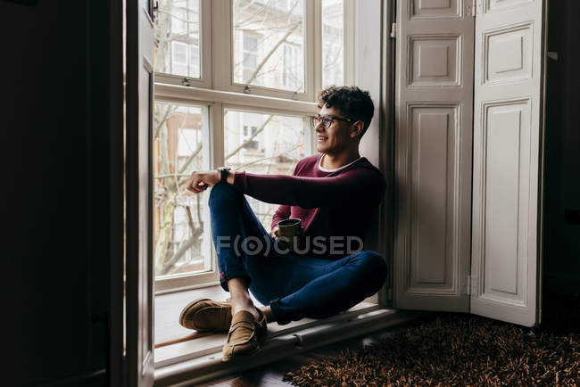 Улыбающийся мужчина с чашкой сидя на подоконнике — стоковое фото