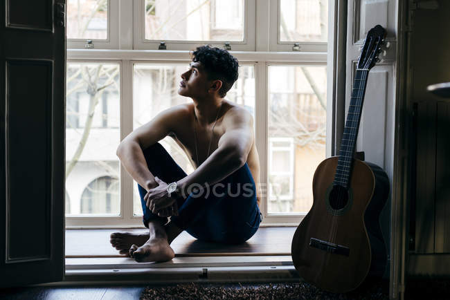 Nachdenklicher Mann ohne Hemd sitzt mit überkreuzten Beinen auf Fensterbank — Stockfoto